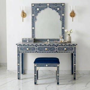 May include: A navy blue vanity set with a matching mirror, stool, and decorative floral inlay. The vanity has three drawers with gold-colored knobs. The mirror has an arched top. A small box and a tray with perfume bottles are on the vanity.