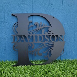 May include: Black metal monogram letter "D" with the name "DAVIDSON" cut out in the center. The letter features decorative flourishes and is mounted against a blue background. The monogram is likely for home decor.