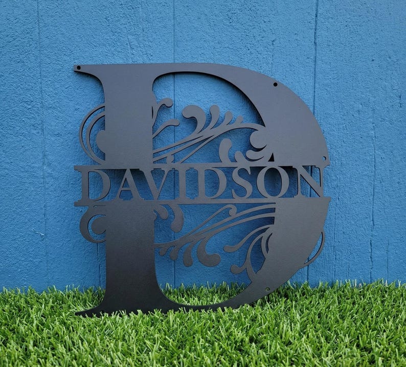 May include: Black metal monogram letter "D" with the name "DAVIDSON" cut out in the center. The letter features decorative flourishes and is mounted against a blue background. The monogram is likely for home decor.