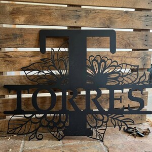 May include: Black metal family name sign with the name "Torres" and the letter "T" in a decorative floral design. The sign is mounted on a wooden pallet background.