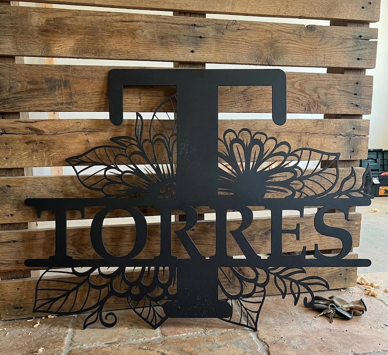 May include: Black metal family name sign with the name "Torres" and the letter "T" in a decorative floral design. The sign is mounted on a wooden pallet background.