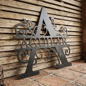 May include: A black metal monogram sign featuring the letter "A" with the name "AGUILAR" cut out in the center. The sign has decorative flourishes and is mounted on a wooden background.