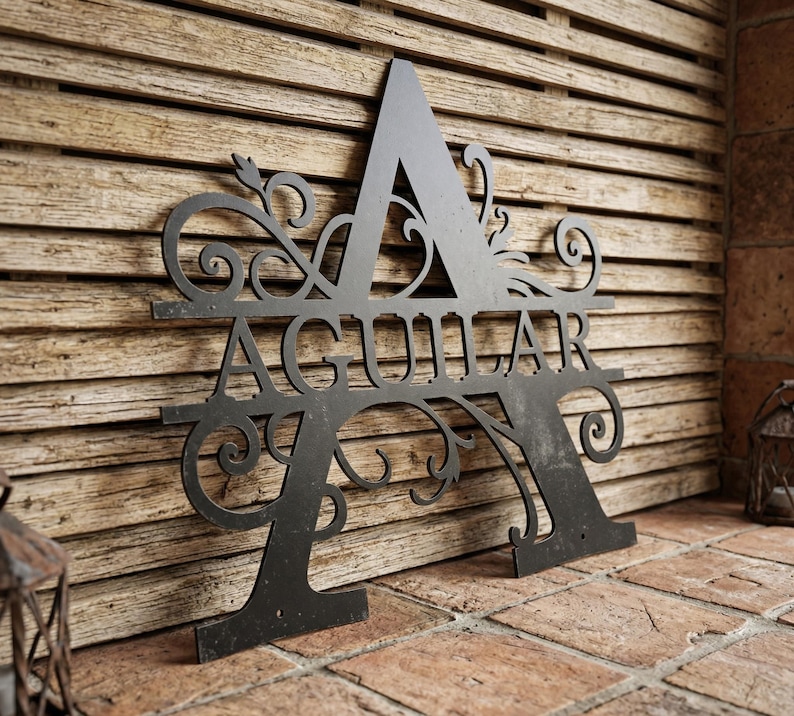 May include: A black metal monogram sign featuring the letter "A" with the name "AGUILAR" cut out in the center. The sign has decorative flourishes and is mounted on a wooden background.