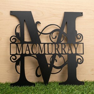 May include: Black metal monogram featuring the letter "M" with the name "MACMURRAY" cut out in the center. The design includes decorative flourishes and is suitable for outdoor or indoor display. The monogram is displayed on a wooden surface.