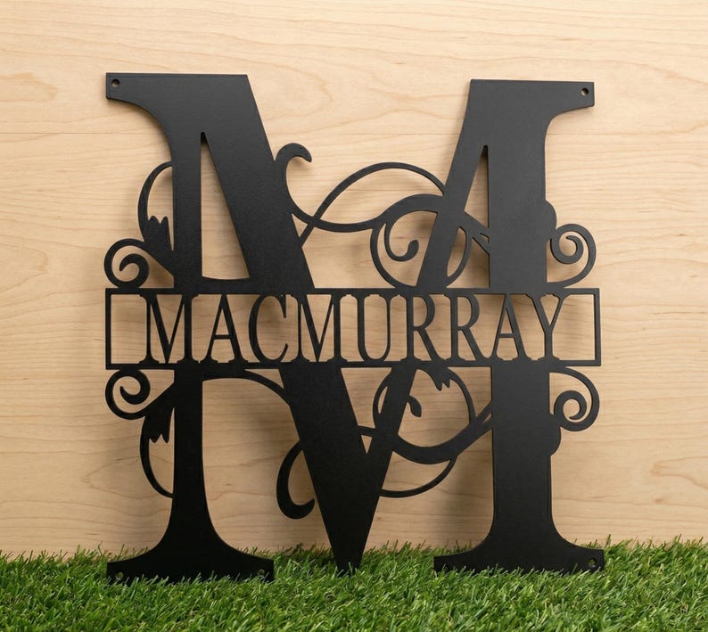 May include: Black metal monogram featuring the letter "M" with the name "MACMURRAY" cut out in the center. The design includes decorative flourishes and is suitable for outdoor or indoor display. The monogram is displayed on a wooden surface.