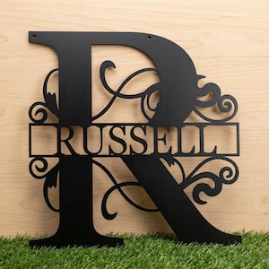 May include: Black metal monogram sign featuring the letter "R" with the name "RUSSELL" cut out. The letter is adorned with decorative flourishes. The sign is displayed against a light wood background and green grass.