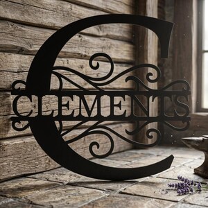 May include: Black metal sign in the shape of the letter "C" with the name "CLEMENTS" cut out in the center. The sign has decorative scrollwork and is displayed against a rustic wooden background.