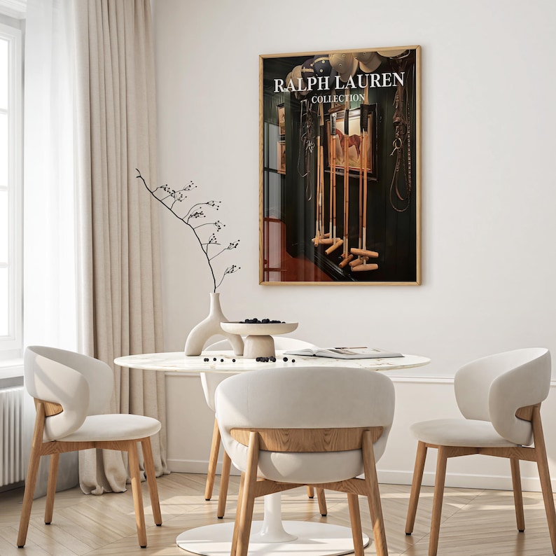 Op de afbeelding: Een ingelijste print met de tekst "RALPH LAUREN COLLECTION" hangt aan een witte muur. De print toont een donker interieur met croquetknuppels en paardensportuitrusting. Een ronde tafel en stoelen staan op de voorgrond.