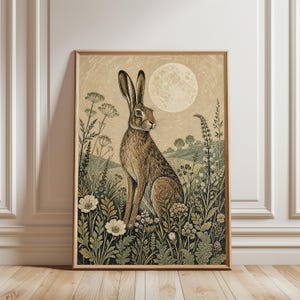May include: A framed wall art print featuring a brown hare sitting in a field of wildflowers under a large full moon. The illustration uses a muted color palette of beige, sage green, and dark brown, depicting a landscape with rolling hills.
