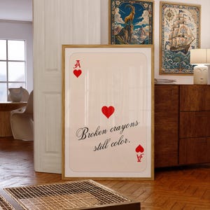 Op de afbeelding: Een rechthoekig ingelijst kunstwerk aan de muur, ontworpen als een extra grote harten aas speelkaart. De gebroken witte kaart heeft rode harten in de hoeken en het midden, met de zwarte cursieve tekst "Broken crayons still color." in het midden.