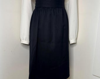 Pode incluir: Um conjunto de duas peças com uma blusa branca de manga comprida e uma saia azul marinho. A blusa tem um decote alto e a saia tem uma cintura larga. A saia vai até à panturrilha.