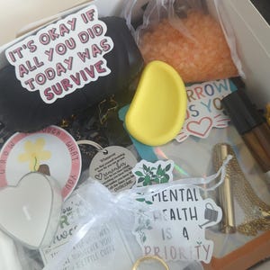 Może przedstawiać: Pudełko prezentowe wypełnione artykułami do pielęgnacji. Zawiera czarny przedmiot z napisem "IT'S OKAY IF ALL YOU DID TODAY WAS SURVIVE", żółty masażer do twarzy, świecę, sole do kąpieli, pierścionek i naszyjnik. Pudełko zawiera również naklejki i małą butelkę.