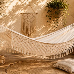 May include: An off-white fabric hammock featuring intricate macrame side panels with decorative fringe tassels. The hammock is suspended by thick woven ropes at both ends, showcasing a detailed knotted pattern along the lower edge.