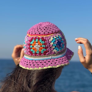 Può includere: Un cappello a secchiello all'uncinetto rosa con motivi quadrati colorati. Il cappello ha un'ampia tesa ed è decorato con quadrati nelle tonalità dell'arancione, del blu e del verde. Un accessorio elegante per le giornate di sole.