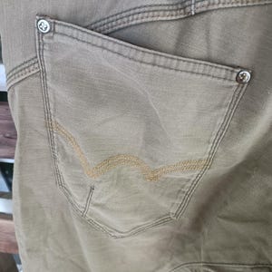 May include: Close-up of khaki-coloured trousers with a back pocket. The pocket has a decorative stitch design in a lighter tan colour. The trousers have visible stitching and metal button details. The fabric appears to be a durable cotton or canvas material.