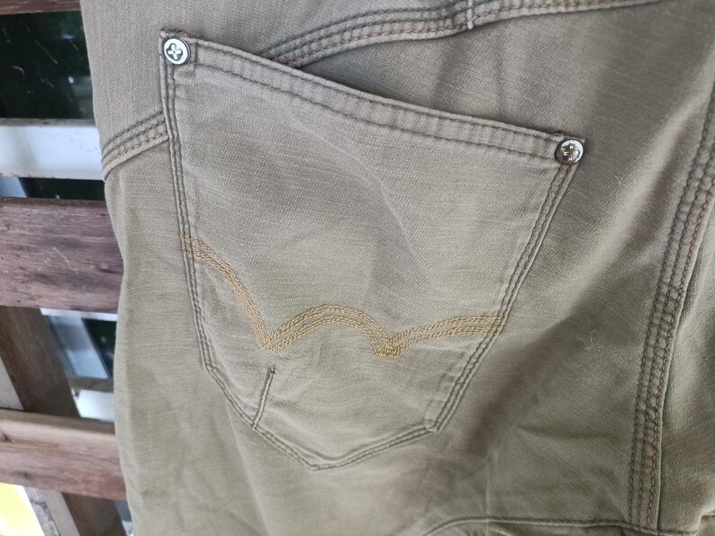 May include: Close-up of khaki-coloured trousers with a back pocket. The pocket has a decorative stitch design in a lighter tan colour. The trousers have visible stitching and metal button details. The fabric appears to be a durable cotton or canvas material.
