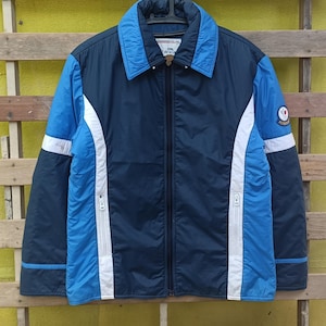 Puede incluir: Chaqueta azul marino y azul claro con cremallera completa. La chaqueta tiene rayas blancas en las mangas y los laterales, y un parche con el logotipo en la manga superior. El cuello es azul claro.