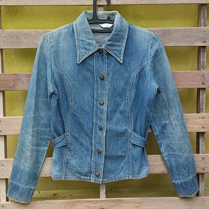May include: A vintage blue denim jacket with a button-up front, collar, and pockets. The jacket is hanging on a black hanger against a wooden background. The denim has a faded appearance.
