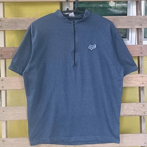 Op de afbeelding: Blauw shirt met korte mouwen met een textuurpatroon en een kwartrits. Het shirt heeft een wit Fox-logo op de borst. Het hangt aan een zwarte hanger tegen een gele en houten achtergrond.