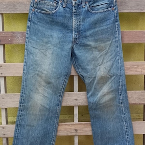 May include: Faded blue denim jeans hanging on a black hanger. The jeans have a button fly, classic five-pocket design, and show signs of wear. The denim is a medium wash with lighter areas.