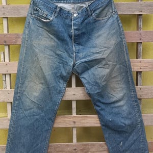 May include: Faded blue denim jeans with a button fly, hanging on a black hanger. The jeans show signs of wear and tear, with visible fading and some minor imperfections. The jeans are a classic style.