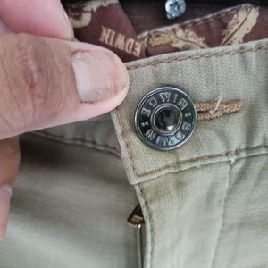 May include: Close-up of khaki trousers featuring a metal button. The button is circular and branded with "EDWIN". A finger is touching the button. The zipper is also visible.