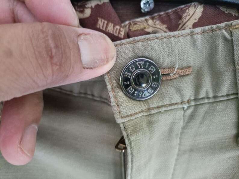 May include: Close-up of khaki trousers featuring a metal button. The button is circular and branded with "EDWIN". A finger is touching the button. The zipper is also visible.