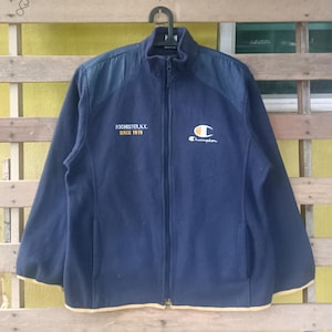 Puede incluir: Chaqueta Champion azul marino con cremallera y cuello alzado. La chaqueta presenta el texto "ROCHESTER N.Y. SINCE 1919" y el logotipo de Champion en blanco y amarillo. La chaqueta tiene un ribete amarillo.