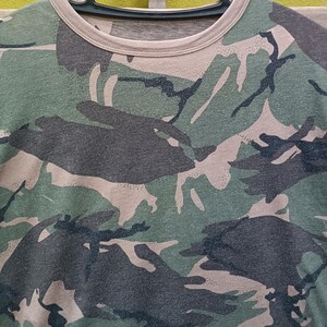 Puede incluir: Camiseta con estampado de camuflaje en tonos verdes, marrones y negros. La camiseta est&aacute; colgada de una percha negra. El dise&ntilde;o de camuflaje cubre toda la camiseta, con cuello redondo y mangas cortas.