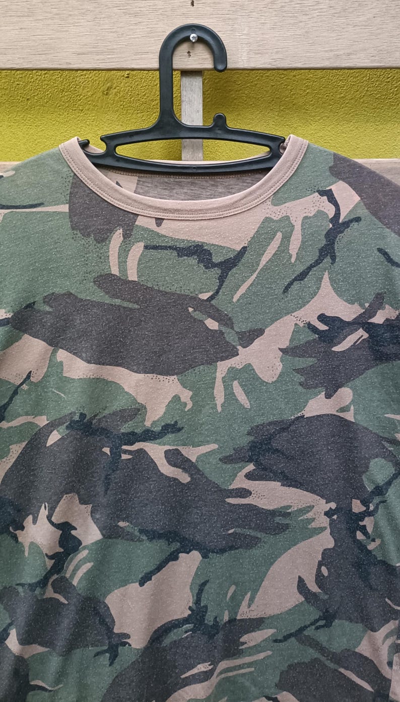 Puede incluir: Camiseta con estampado de camuflaje en tonos verdes, marrones y negros. La camiseta est&aacute; colgada de una percha negra. El dise&ntilde;o de camuflaje cubre toda la camiseta, con cuello redondo y mangas cortas.