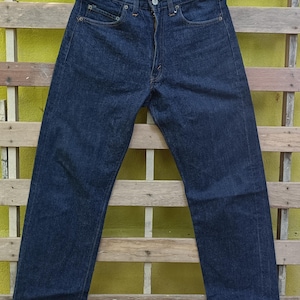 May include: Dark blue denim jeans hanging on a black hanger. The jeans have a classic five-pocket design, a button and zipper closure, and are made of a sturdy denim fabric. The jeans are displayed against a wooden background.