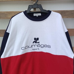 May include: A color-blocked sweatshirt in white, red, and navy blue, featuring the "courr&egrave;ges sport" logo in navy blue. The sweatshirt is hanging on a black hanger against a wooden background.