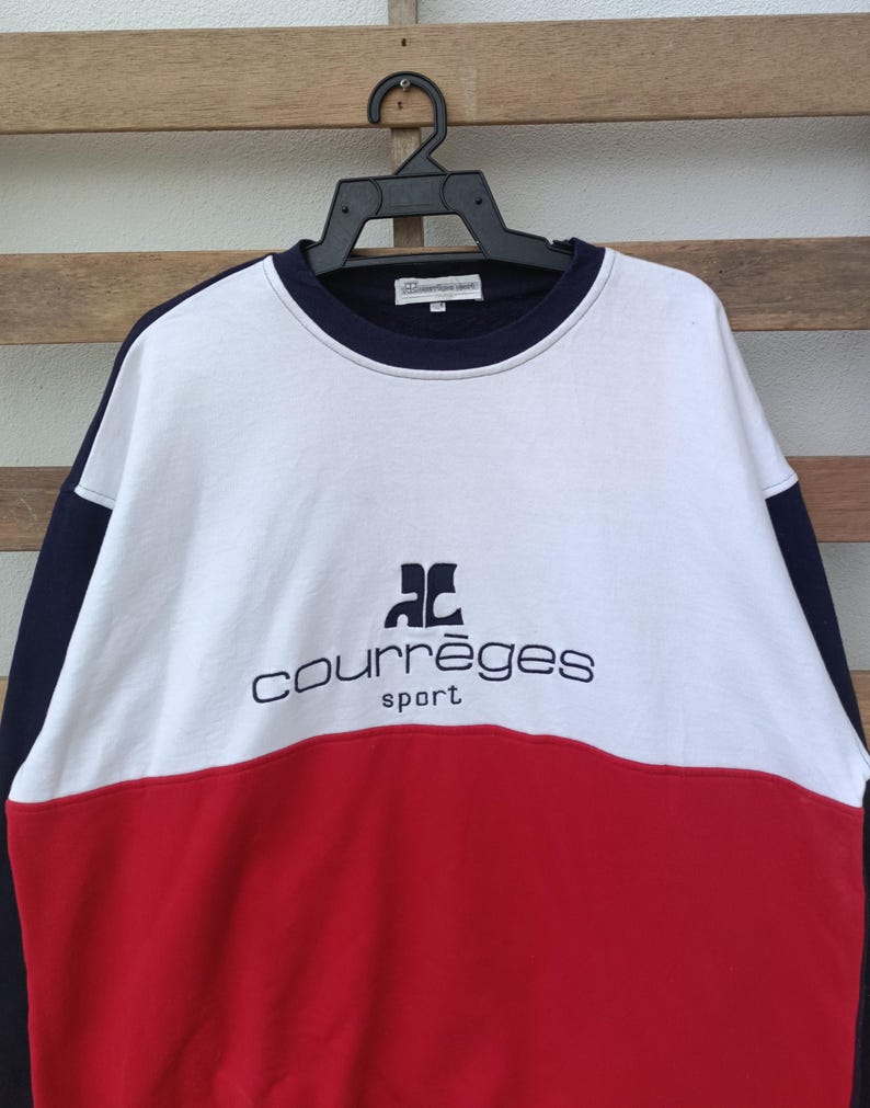 May include: A color-blocked sweatshirt in white, red, and navy blue, featuring the "courr&egrave;ges sport" logo in navy blue. The sweatshirt is hanging on a black hanger against a wooden background.