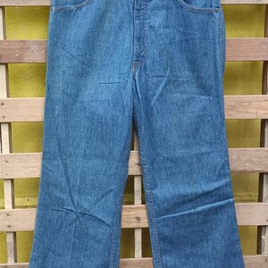 May include: Blue denim jeans with a classic five-pocket design, hanging on a black hanger. The jeans have a straight leg cut and a button and zipper closure. The denim has a medium wash and a slight fade.