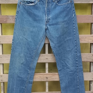 May include: Faded blue denim jeans hanging on a black hanger. The jeans have a classic five-pocket design, a button fly, and show signs of wear and tear, with some fading and distressing. The jeans are hanging against a wooden background.