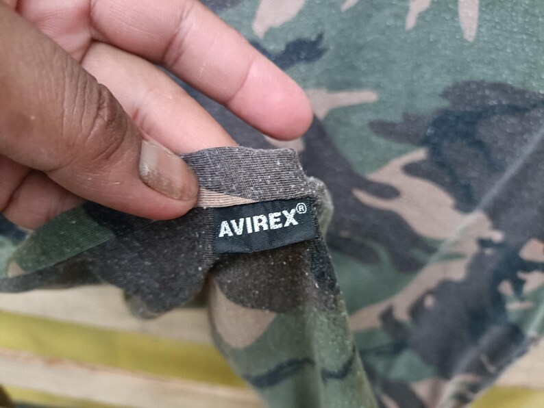 Puede incluir: Primer plano de una tela con estampado de camuflaje con la marca "AVIREX&reg;" impresa en una etiqueta negra. La tela presenta una mezcla de colores verdes, marrones y beiges.