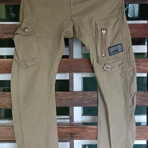 May include: Khaki cargo trousers with multiple pockets and a zip pocket on the right leg. The trousers have a button and zip closure, belt loops, and decorative stitching. The trousers are hanging on a wooden background.