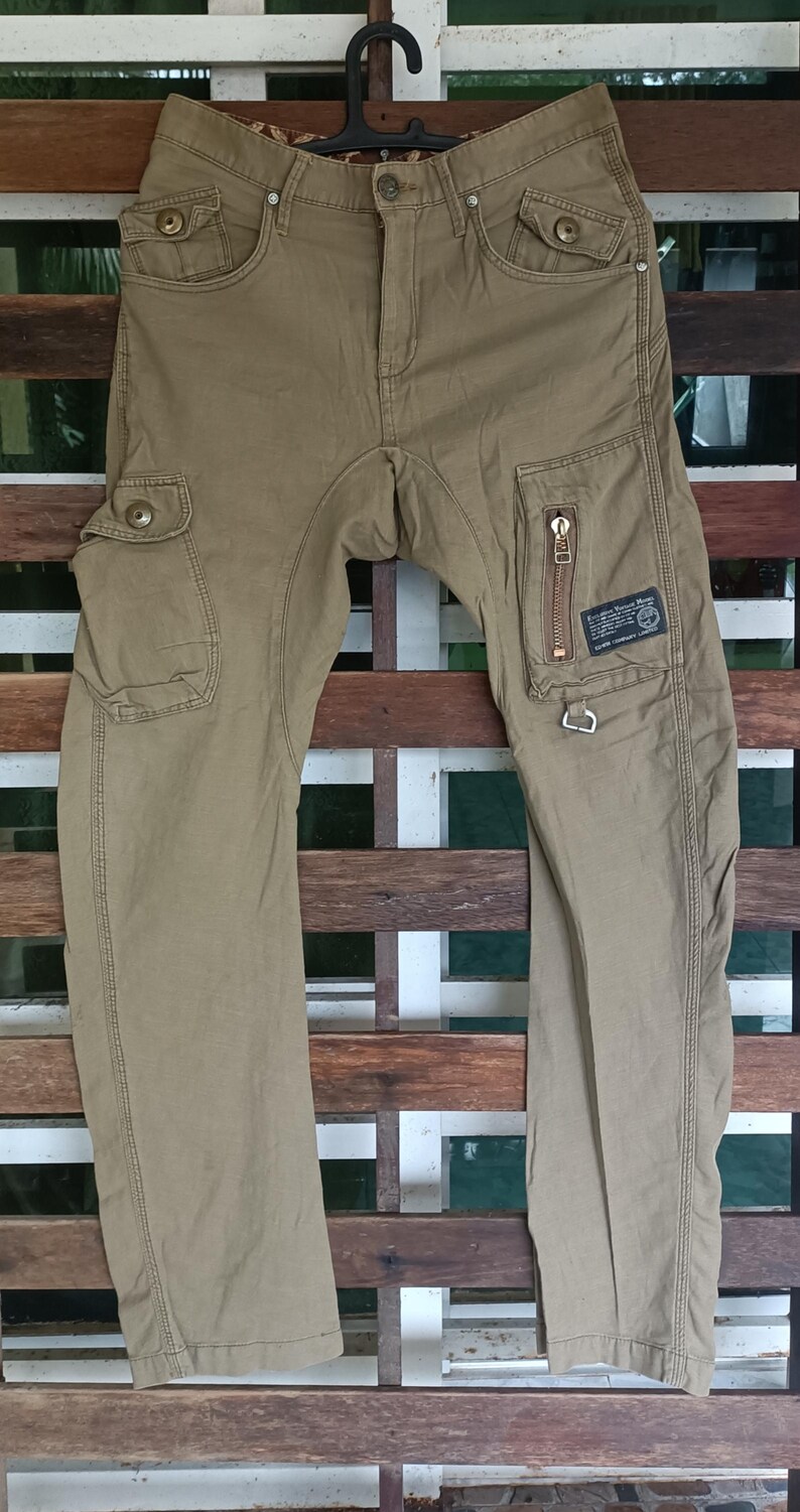 May include: Khaki cargo trousers with multiple pockets and a zip pocket on the right leg. The trousers have a button and zip closure, belt loops, and decorative stitching. The trousers are hanging on a wooden background.