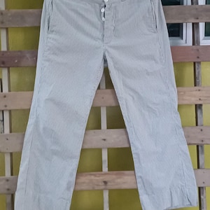 Puede incluir: Pantalones a rayas grises y blancas claras colgados de una percha negra. Los pantalones tienen cierre de botón y bolsillos. La tela tiene una textura de seersucker. Los pantalones se muestran sobre un fondo de madera.