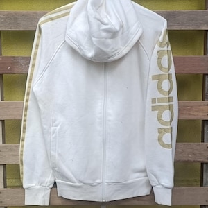 May include: White zip-up hoodie with a hood and gold stripes on the sleeves. The word "adidas" is printed in gold on the left sleeve. The hoodie is made of a soft, comfortable material.