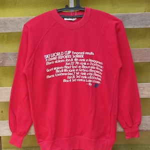 Peut inclure: Sweat-shirt rouge avec texte blanc détaillant les résultats de courses de ski, dont "SKI WORLD CUP" et "P.MAHRE 3rdRANK". Le texte mentionne des lieux comme Renggries, Chamonix et Lake Louise. Le sweat-shirt a des manches longues et un col rond.