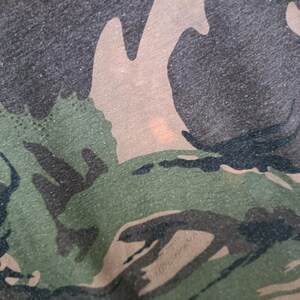 Puede incluir: Primer plano de una tela con estampado de camuflaje. El dise&ntilde;o presenta una mezcla de formas verdes, marrones y negras, creando una apariencia texturizada. La tela tiene un aspecto ligeramente desgastado, con sutiles variaciones de color y textura.