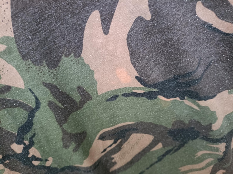 Puede incluir: Primer plano de una tela con estampado de camuflaje. El dise&ntilde;o presenta una mezcla de formas verdes, marrones y negras, creando una apariencia texturizada. La tela tiene un aspecto ligeramente desgastado, con sutiles variaciones de color y textura.