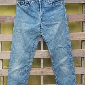 May include: Light blue denim jeans hanging on a black hanger. The jeans have a classic five-pocket design, a button and zipper closure, and show signs of wear. The jeans are displayed against a wooden background.