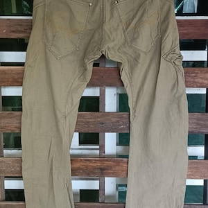 May include: Khaki-coloured trousers with a classic design, featuring two back pockets with decorative stitching and a belt loop. The trousers are hanging on a black hanger against a wooden background. The trousers appear to be made of a durable fabric.