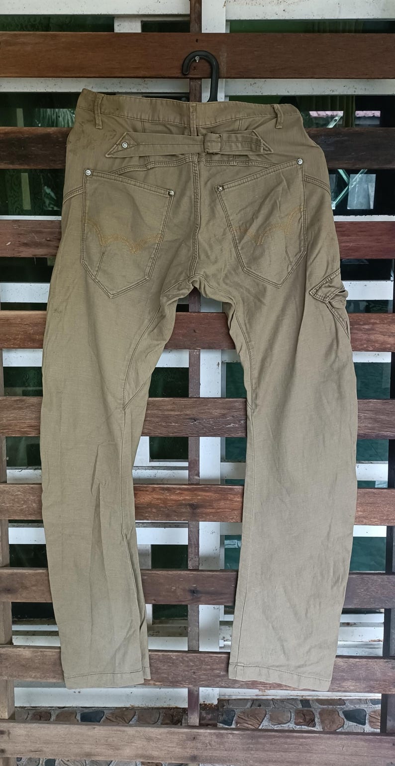 May include: Khaki-coloured trousers with a classic design, featuring two back pockets with decorative stitching and a belt loop. The trousers are hanging on a black hanger against a wooden background. The trousers appear to be made of a durable fabric.