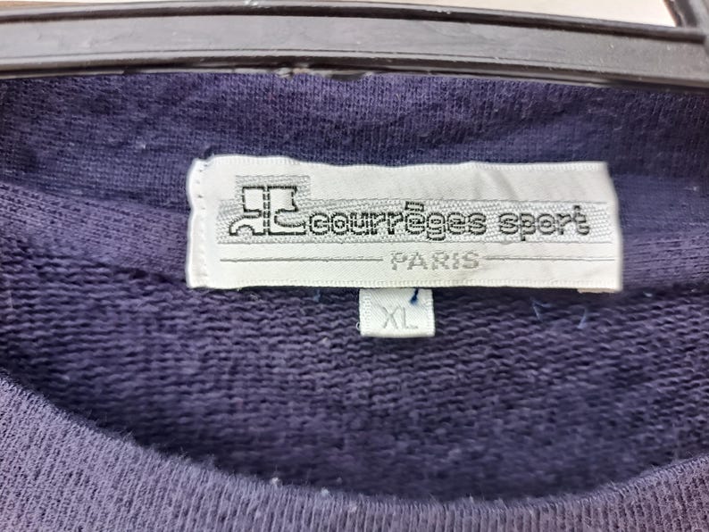 May include: Close-up of a navy blue sweatshirt with a white tag. The tag reads "courr&egrave;ges sport PARIS" with an XL size label. The sweatshirt is hanging on a black hanger.