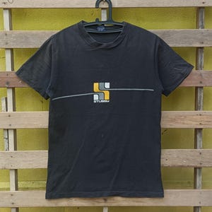 May include: Black short-sleeved t-shirt with a horizontal grey line and a logo in yellow, white, and orange. The logo features the word "STUSSY" in white. The shirt is hanging on a black hanger.