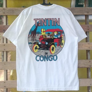 May include: White t-shirt featuring a colorful graphic from the Tintin in Congo comic. The design includes a vintage car, a giraffe, and the text "TINTIN IN CONGO" in red and blue.