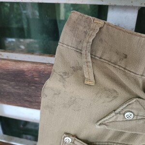 May include: Close-up of beige trousers showing wear and tear. The image focuses on the waistband, belt loops, and a pocket with a decorative button. The fabric shows signs of use, with stains and fading.